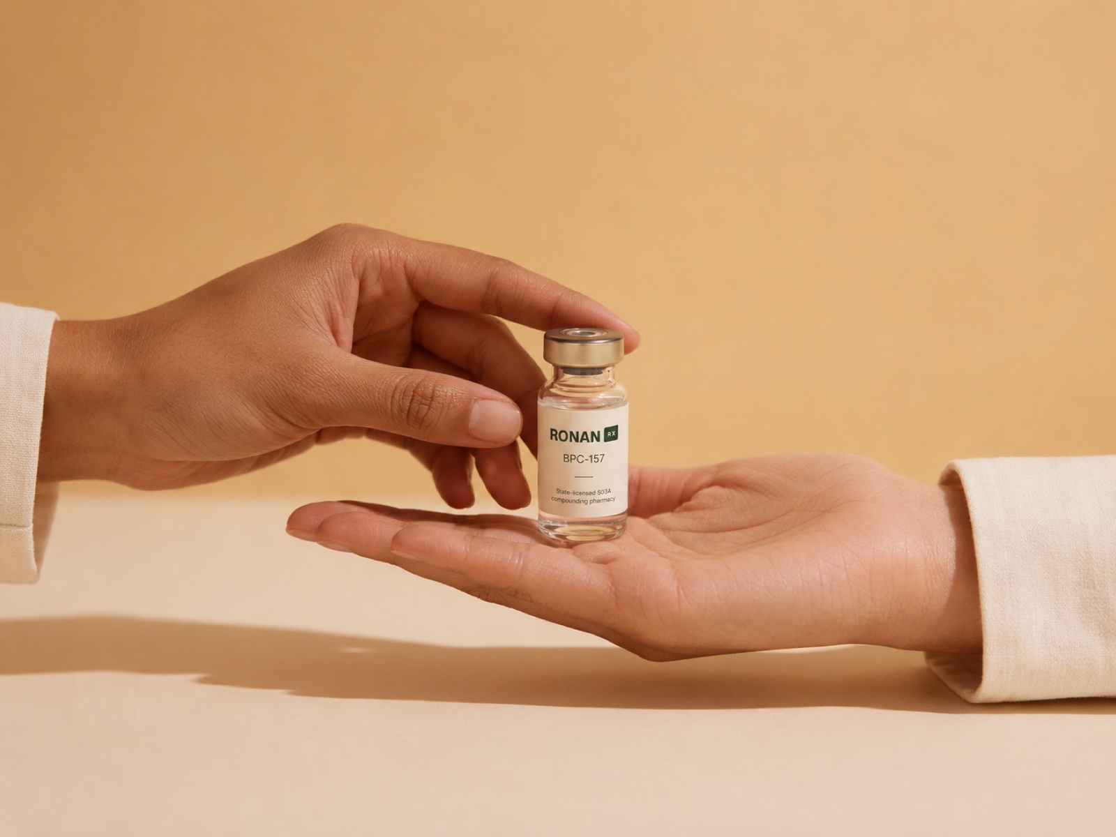 One clinician's hand offering a labeled RonanRx BPC-157 vial to a second clinician's open palm, against a warm cream background.